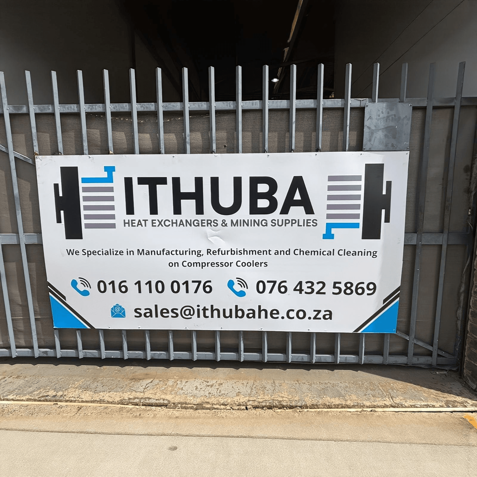Sign on a metal gate reads 'Ithuba Heat Exchangers & Mining Supplies'. It includes contact details and services like manufacturing and refurbishment.