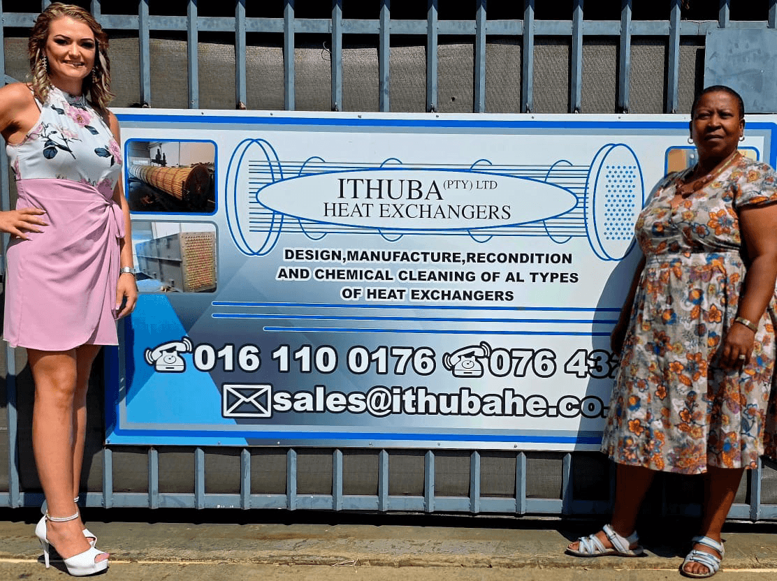 The two directors stand beside a historical sign for "Ithuba Heat Exchangers" on a sunny day. The sign details services and contact information. Both appear relaxed and confident.