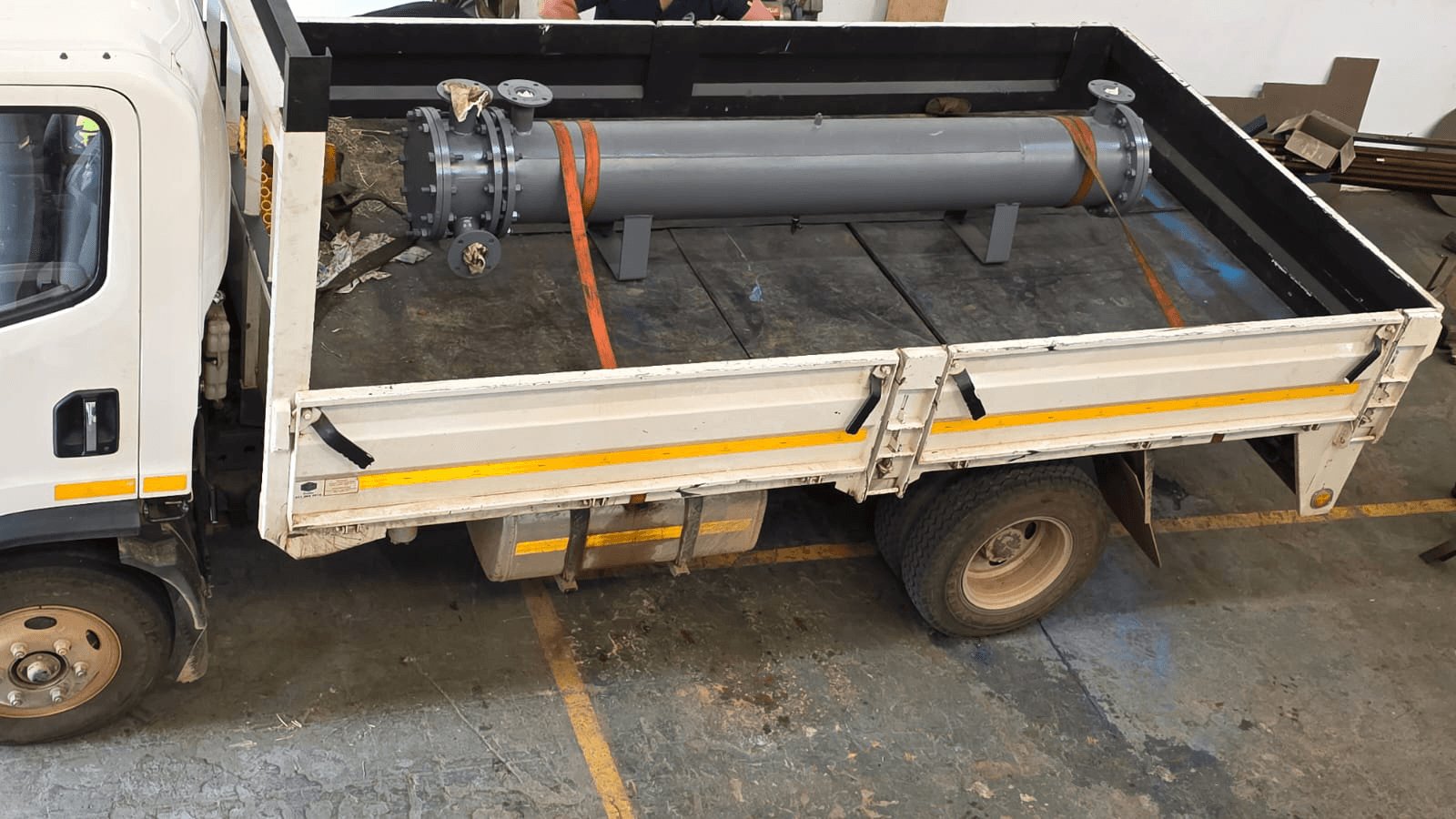 A white flatbed truck parked indoors, carrying an heat exchanger, ready to be shipped, heat exchanger object secured by orange straps. The setting is industrial, with a utilitarian feel.