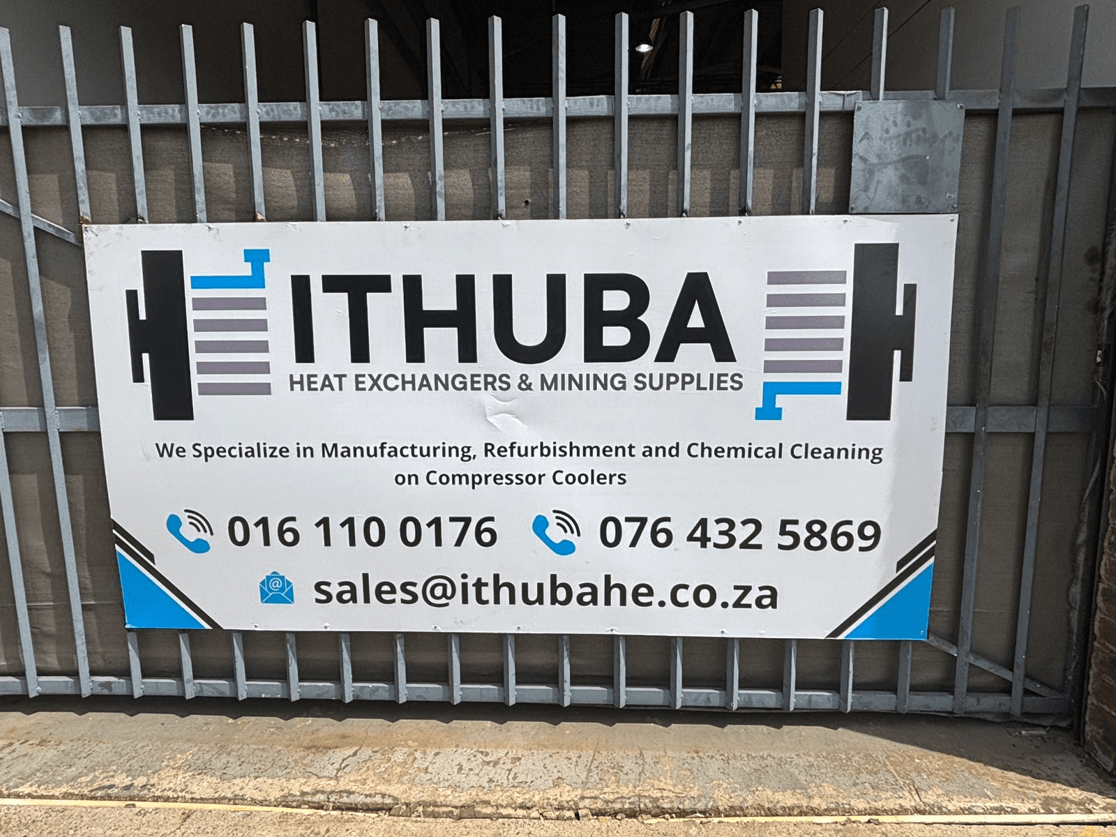 Sign on a metal gate for Ithuba Heat Exchangers & Mining Supplies. It lists contacts and services like manufacturing and cleaning compressor coolers.