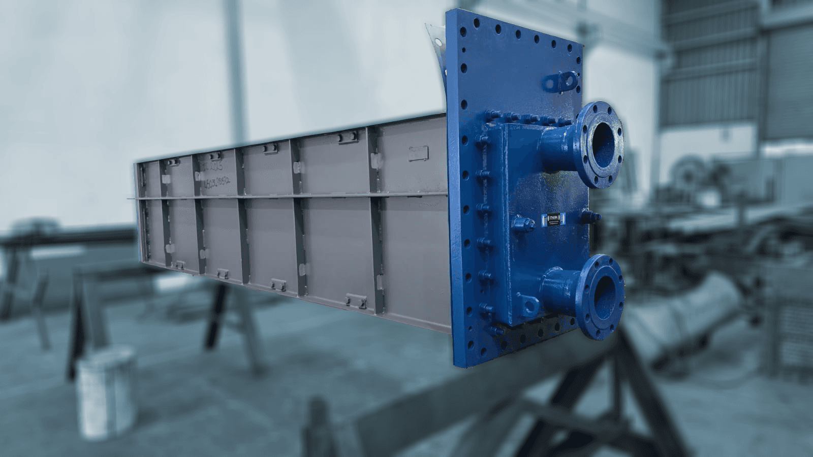 A large industrial heat exchanger with a blue front featuring flanges and bolts stands prominently in a blurred workshop, conveying an engineering focus.