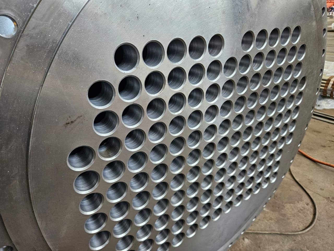 Close-up of a metal tube sheet with evenly spaced circular holes. The shiny metallic surface reflects the industrial setting and precise engineering.